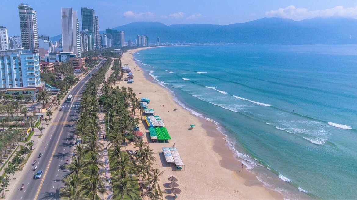 A panoramic aerial view of a long sandy shoreline with turquoise water and the city skyline in the background.