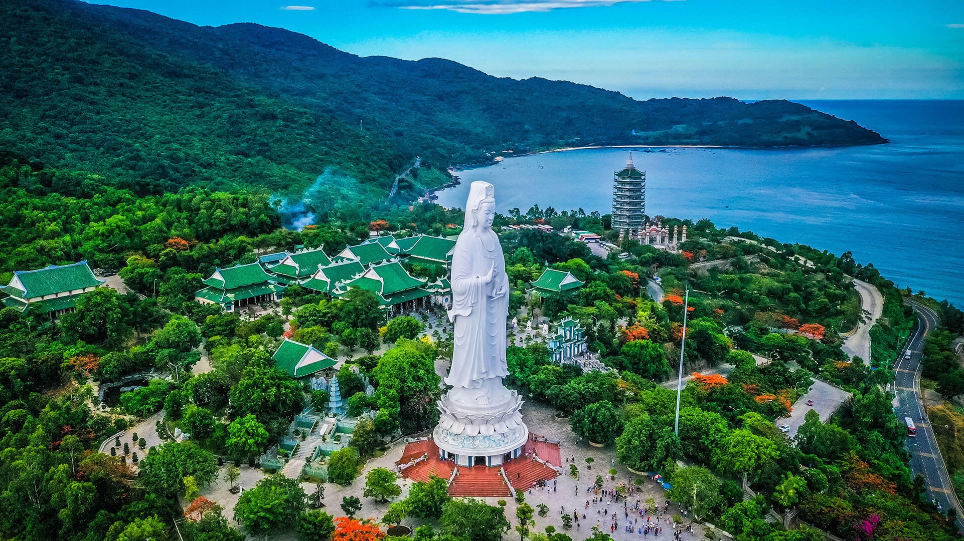 Towering white Lady Buddha statue overlooking the coastline from a hilltop pagoda.